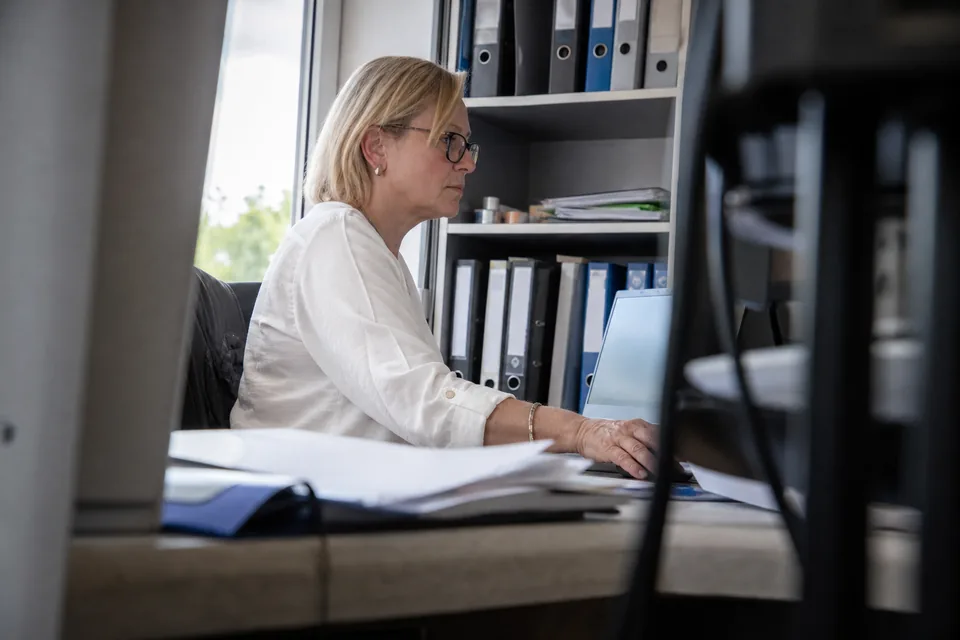 Kobieta w biurze sprawdza dane kontrahenta na laptopie, w tle segregatory