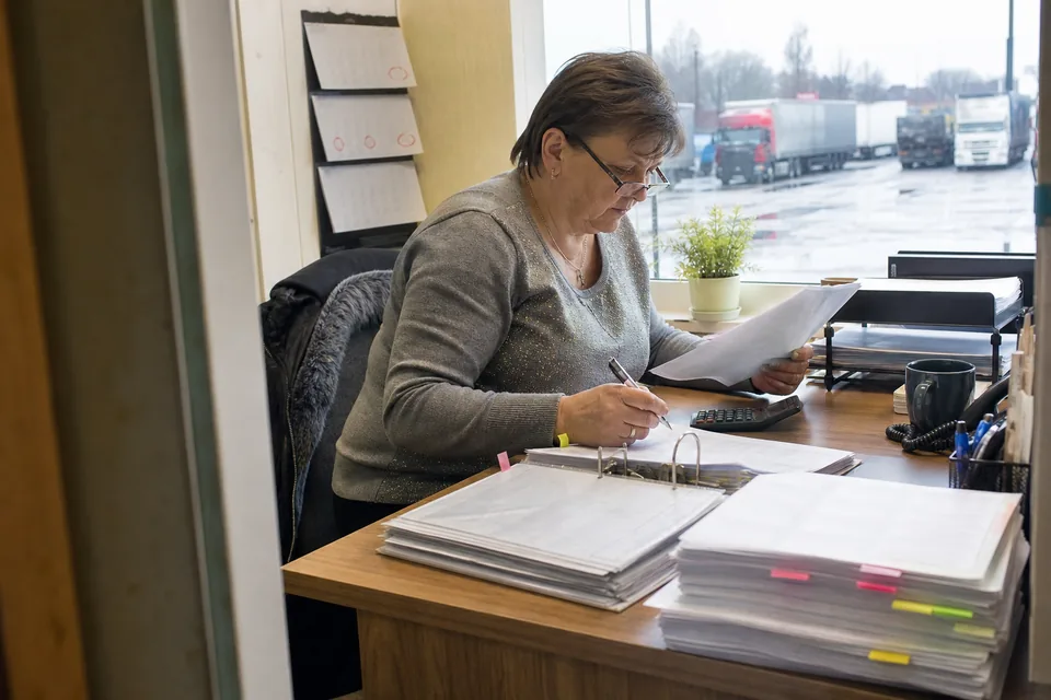 Księgowa firmy przewozowej porównuje listę zleceń z wpłatami od spedycji w małym biurze przy bazie transportowej